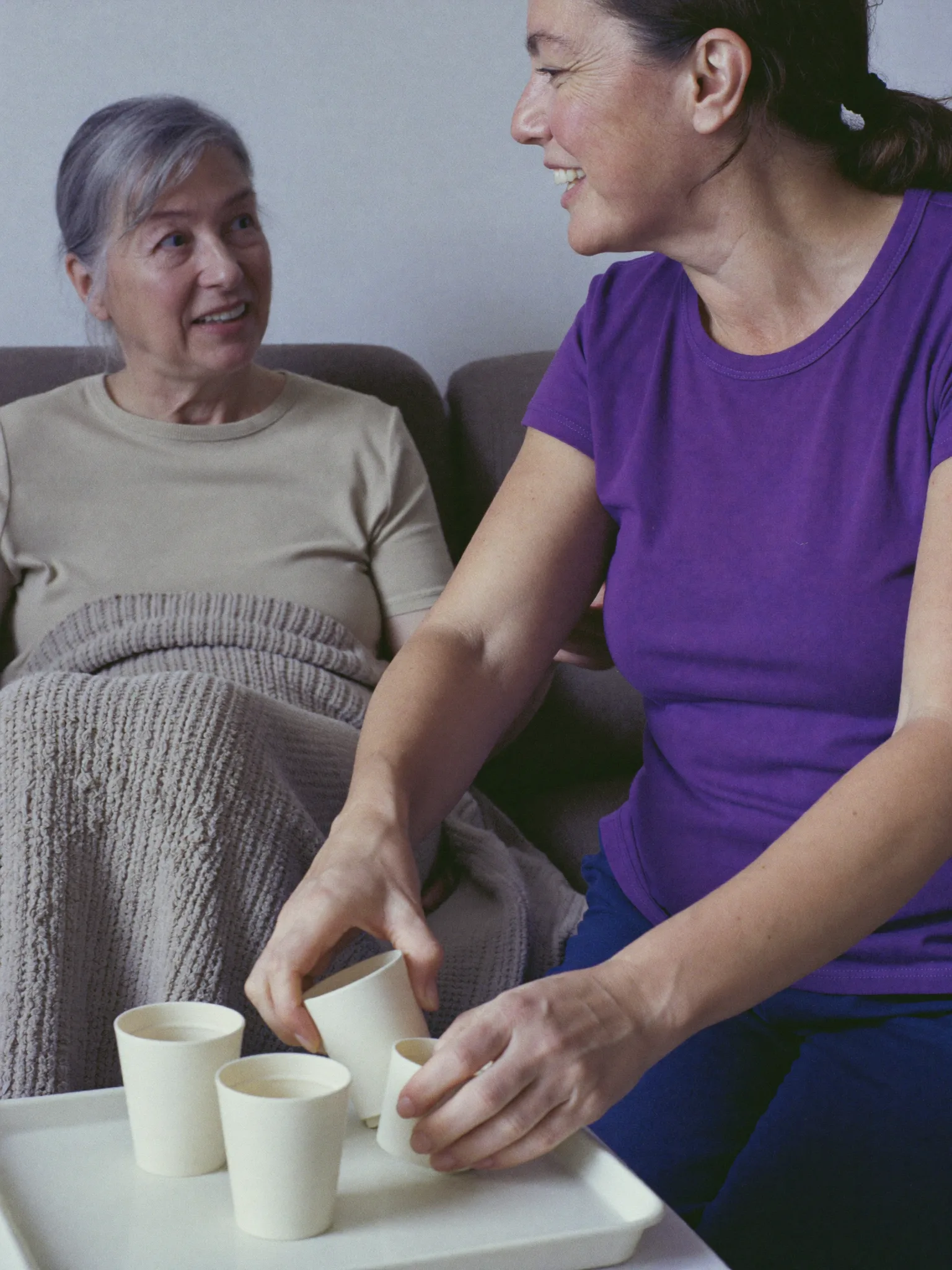 Haushaltshilfe stellt Becher auf den Tisch, eine Seniorin ruht unter einer Decke